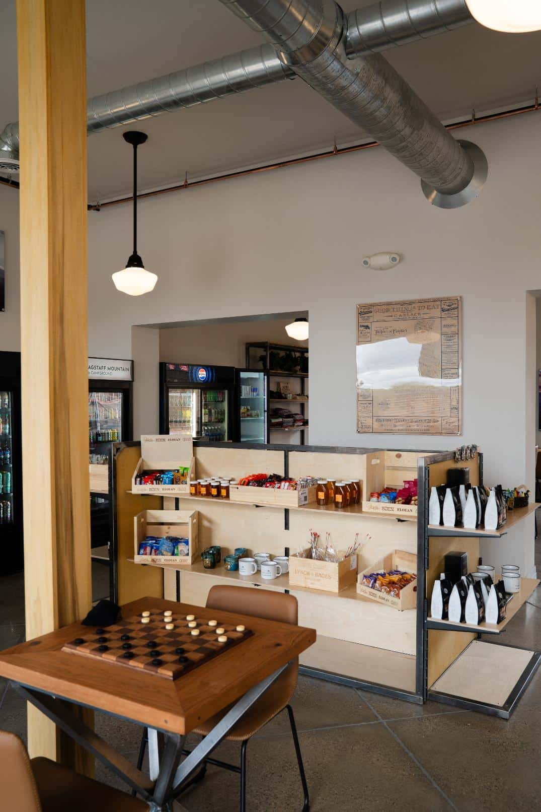 A casual indoor space featuring a wooden checkers table in the foreground and retail shelves stocked with snacks, mugs, and coffee bags in the background.