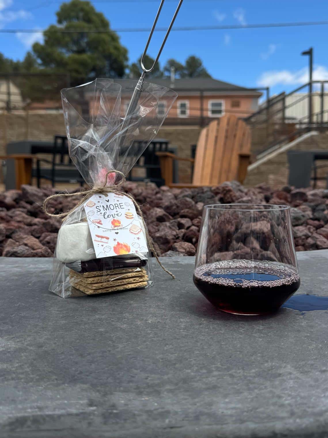 A packaged s'mores kit and a stemless glass of red wine sitting on the edge of an outdoor stone fire pit.