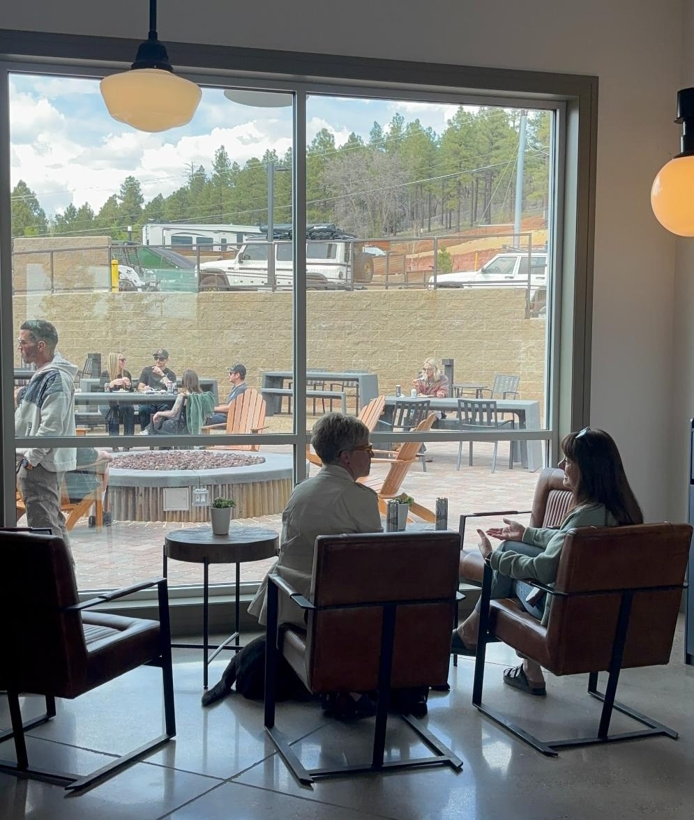 Two women sitting in brown leather chairs indoors, talking near a large floor-to-ceiling window that looks out onto a patio with a fire pit and pine trees.